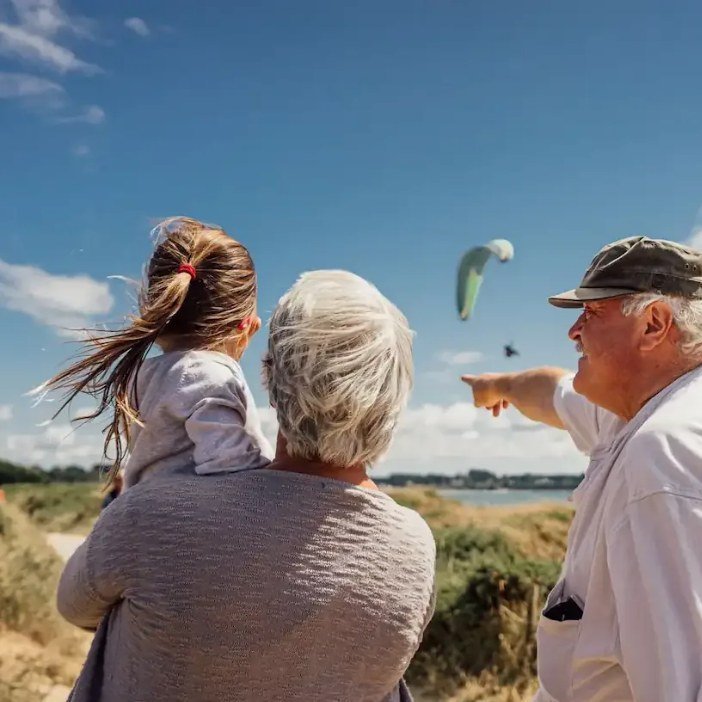 Morföräldrar med ett barnbarn på axlarna tittar på en skärmflygare vid havet